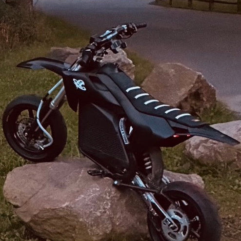 A black electric dirt bike with white graphic stripes on the seat, parked on a large rock on a paved road with trees in the background.